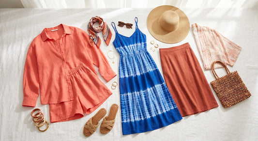 A bright editorial flat lay of a curated summer capsule wardrobe featuring linen coordinates, sundresses, and bold accessories in coral, cobalt, and terracotta tones arranged on a white surface.