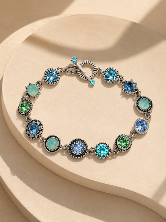 A silver-toned bracelet with alternating round blue and green gemstone settings, displayed on a beige sculptural surface with soft shadows-