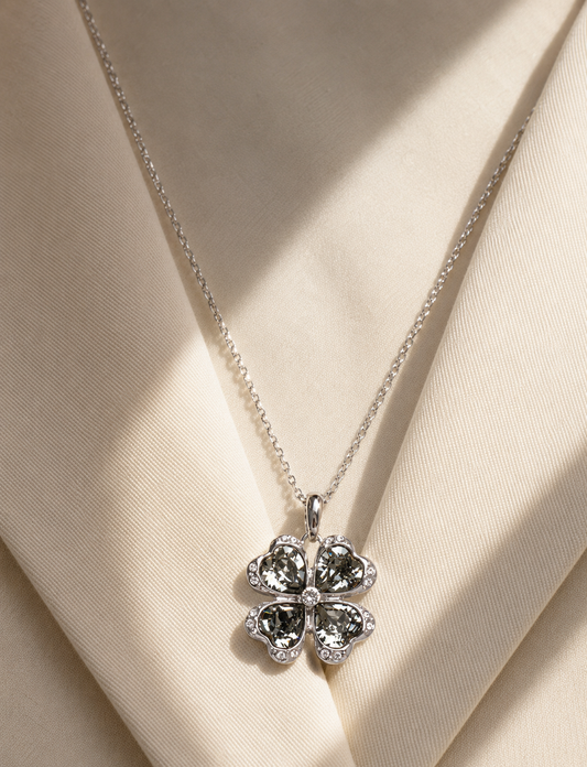 A silver four-leaf clover pendant necklace with clear sparkling stones, displayed on a cream satin fabric background with soft diagonal shadows.