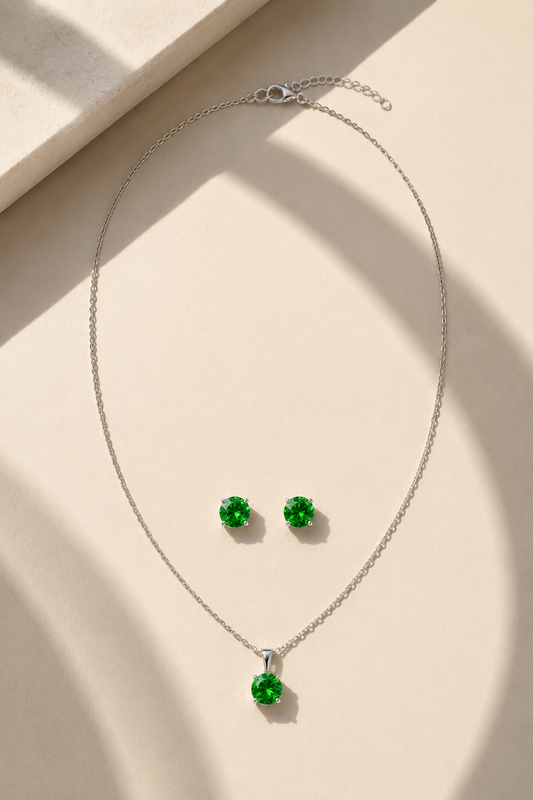 A silver necklace and matching stud earrings set with round green gemstones, arranged on a warm beige surface with soft diagonal shadows.-