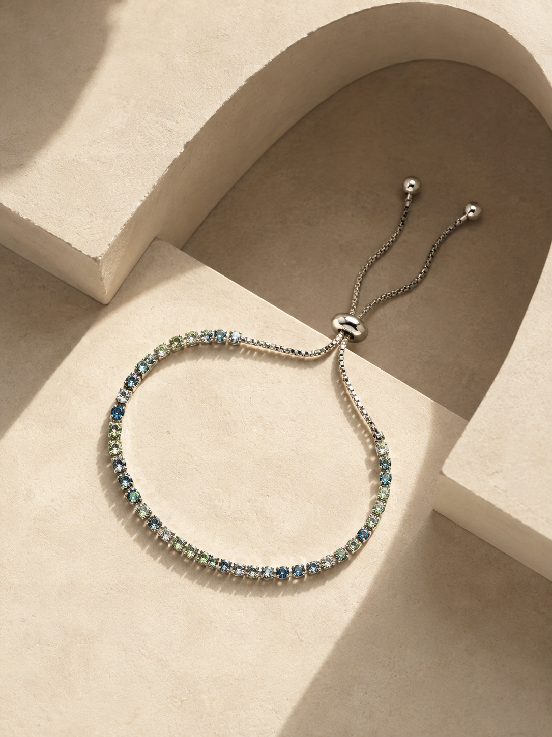 A silver-toned adjustable bracelet with a row of small blue and green iridescent stones, displayed on a beige geometric surface with dramatic shadows.