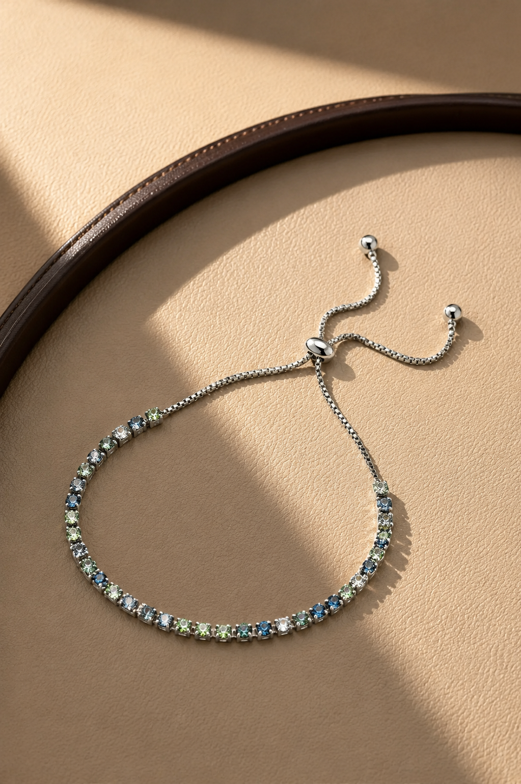 A delicate silver chain bracelet with a row of small blue and green gemstones, displayed on a textured beige surface with a dark brown leather belt edge in the background.-