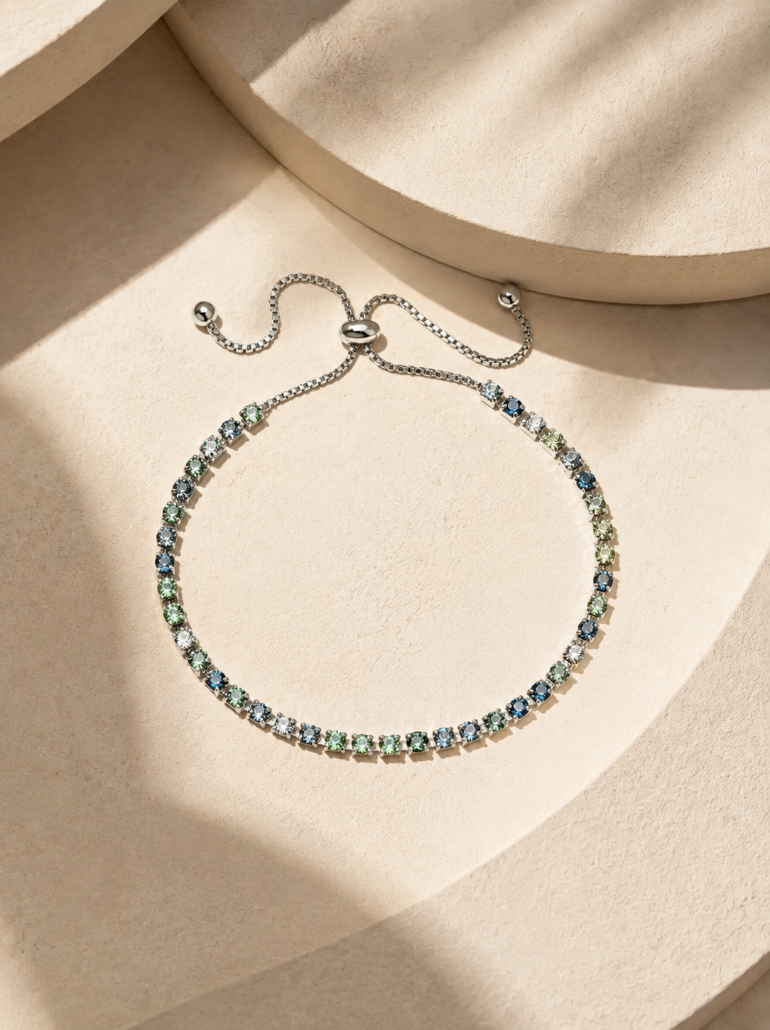 A delicate silver-tone adjustable chain bracelet adorned with small, round, light blue and mint green gemstones, displayed on a beige textured surface with soft shadows.