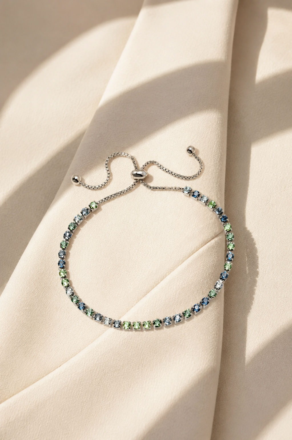 A delicate silver chain bracelet with a row of small iridescent blue-green gemstones, displayed on draped cream-colored fabric with soft shadows.-
