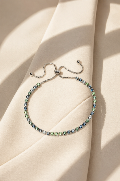 A delicate silver chain bracelet with a row of small iridescent blue-green gemstones, displayed on draped cream-colored fabric with soft shadows.-