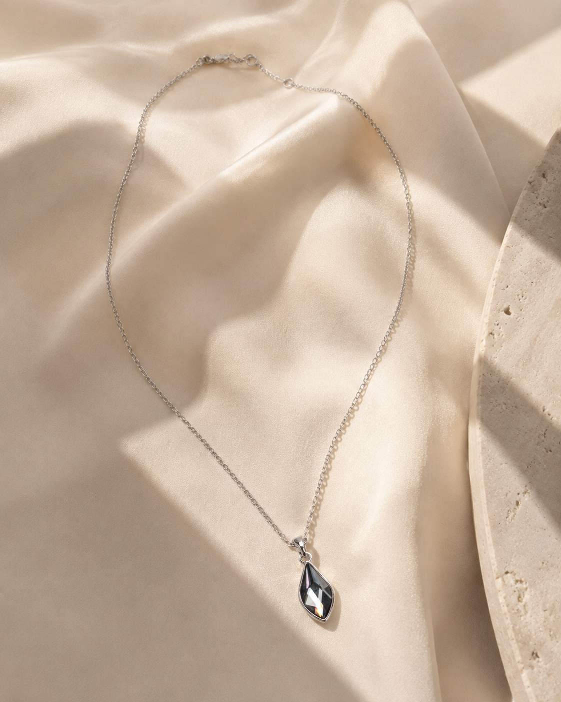 A delicate silver chain necklace with a teardrop-shaped faceted gemstone pendant, displayed on draped cream-colored satin fabric with soft shadows.-