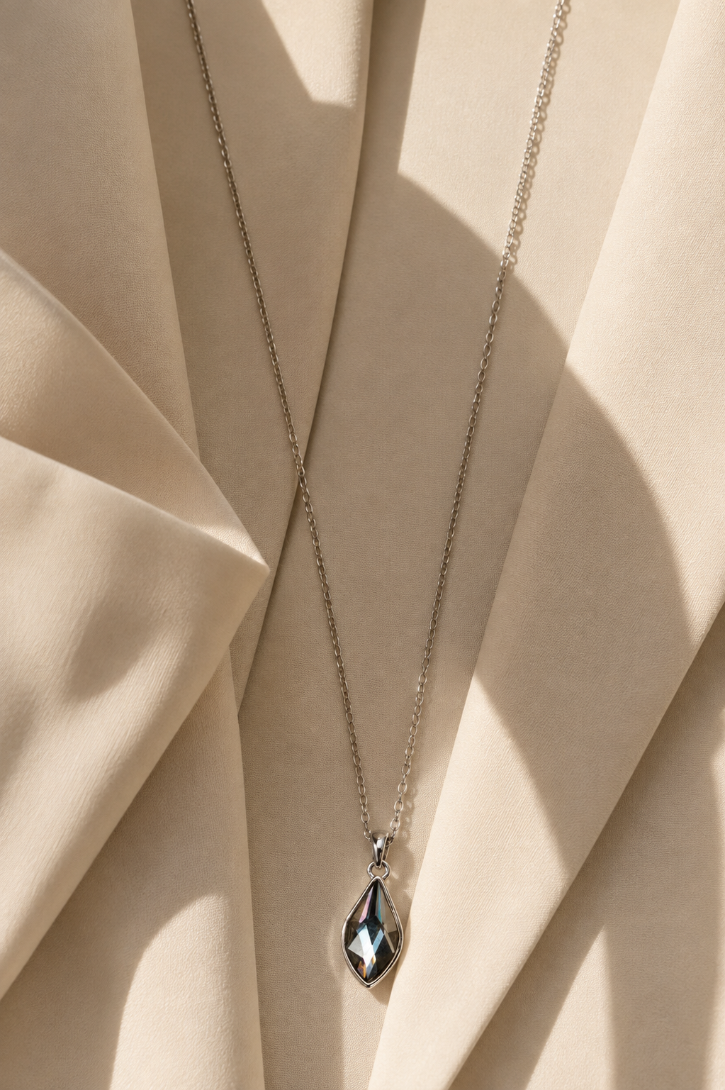 A silver-toned chain necklace with a faceted teardrop pendant, draped over draped beige fabric with soft shadows.