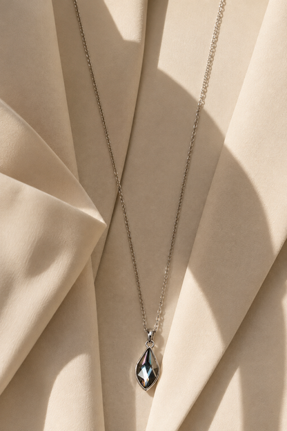 A silver-toned chain necklace with a faceted teardrop pendant, draped over draped beige fabric with soft shadows.