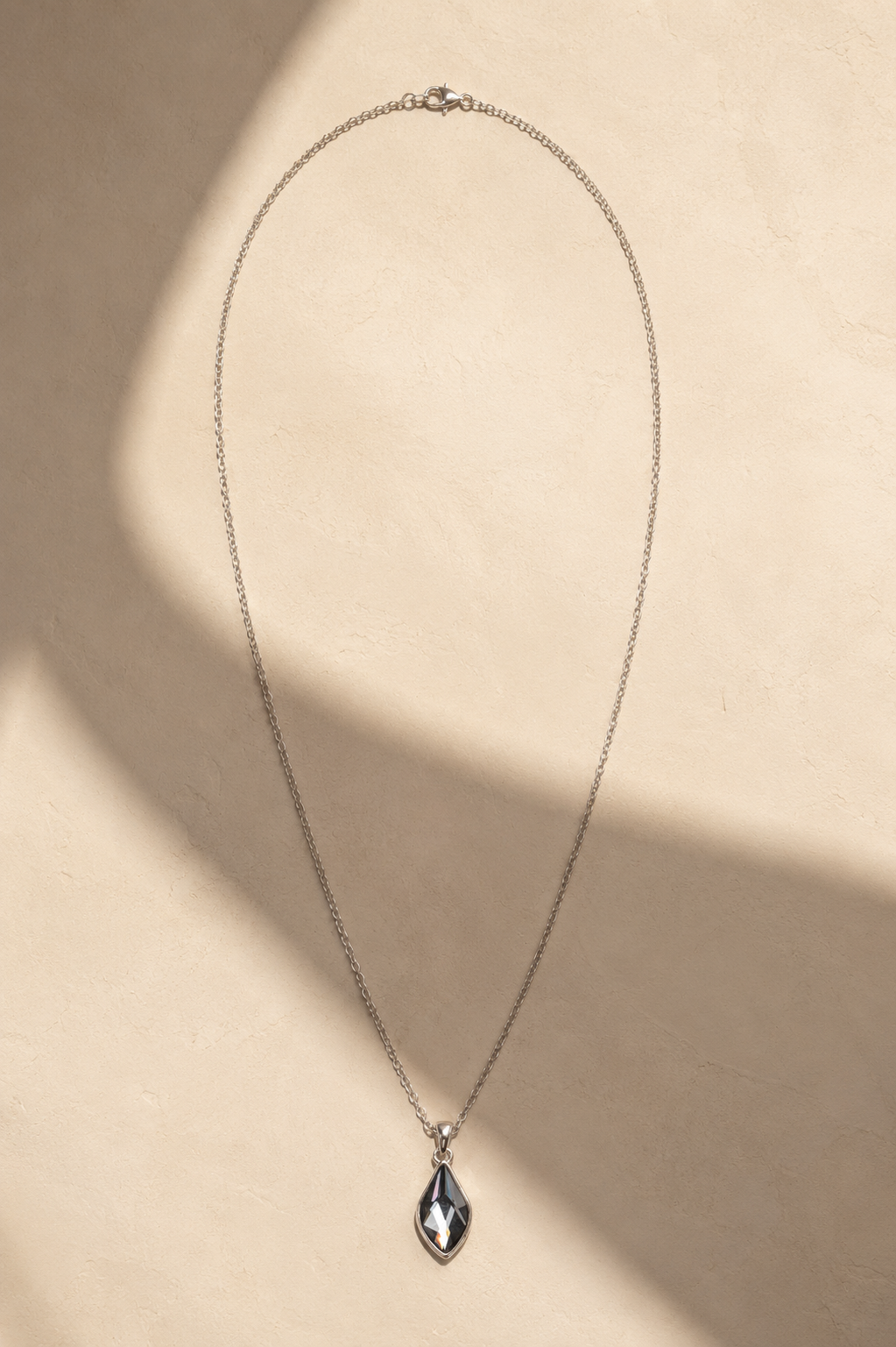 A delicate silver chain necklace with a teardrop-shaped dark gray pendant, displayed on a textured beige background with soft diagonal shadows.-