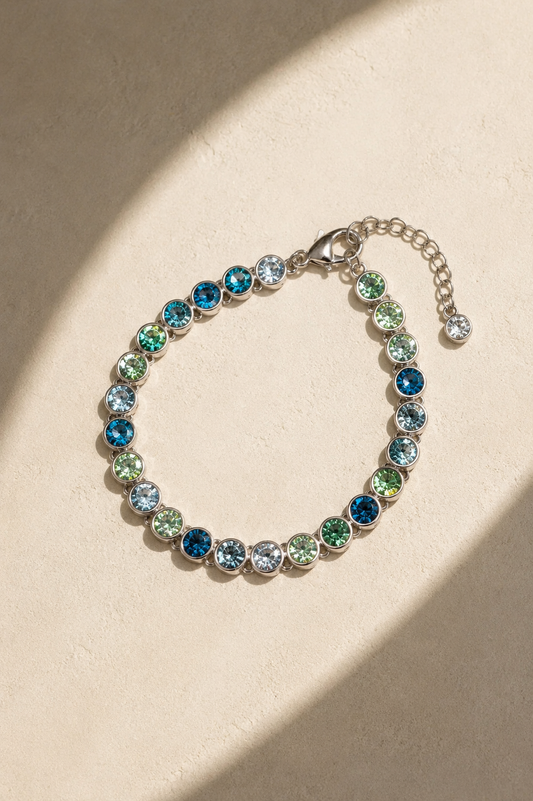 A silver-toned bracelet with round, faceted gemstones in shades of blue, green, and clear, arranged in a circle on a textured beige surface with soft diagonal shadows.-