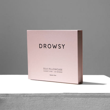 Female model holding a pastel pink coloured Drowsy silk pillowcase box on a white stand with a grey background