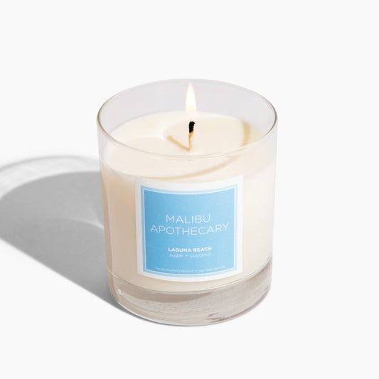 Clear candle with blue label with notes of sugar, coconut and vanilla bean lit up with a white background.